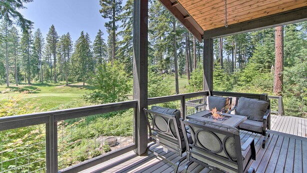 A covered outdoor deck with a fire pit and seating, overlooking a forested area with a golf course in the distance. - Modern Alpine Home on Prospector Golf Course! (Cle Elum)