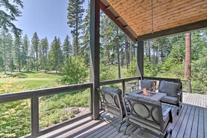 A covered outdoor deck with a fire pit and seating, overlooking a forested area with a golf course in the distance. - Modern Alpine Home on Prospector Golf Course! (Cle Elum)