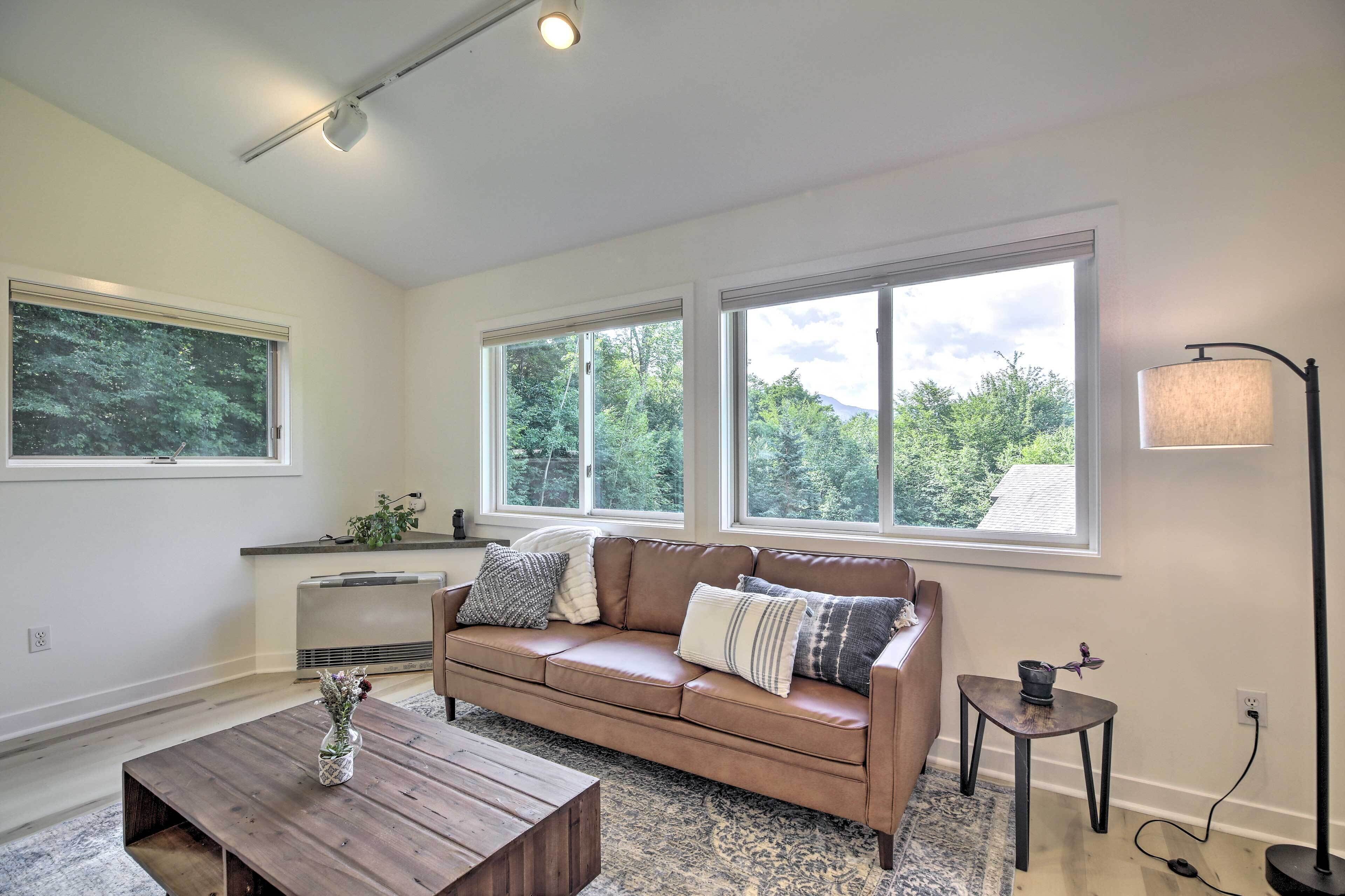 A living room with a brown leather sofa, a wooden coffee table, and a small side table. There are large windows with a view of trees and a lamp on a side table.