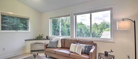 A living room with a brown leather sofa, a wooden coffee table, and a small side table. There are large windows with a view of trees and a lamp on a side table.