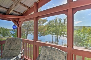 A covered outdoor seating area with wooden railings and chairs, overlooking a lake and trees. - Inviting Lakefront Home: Seasonal Boat Dock! (Holden)