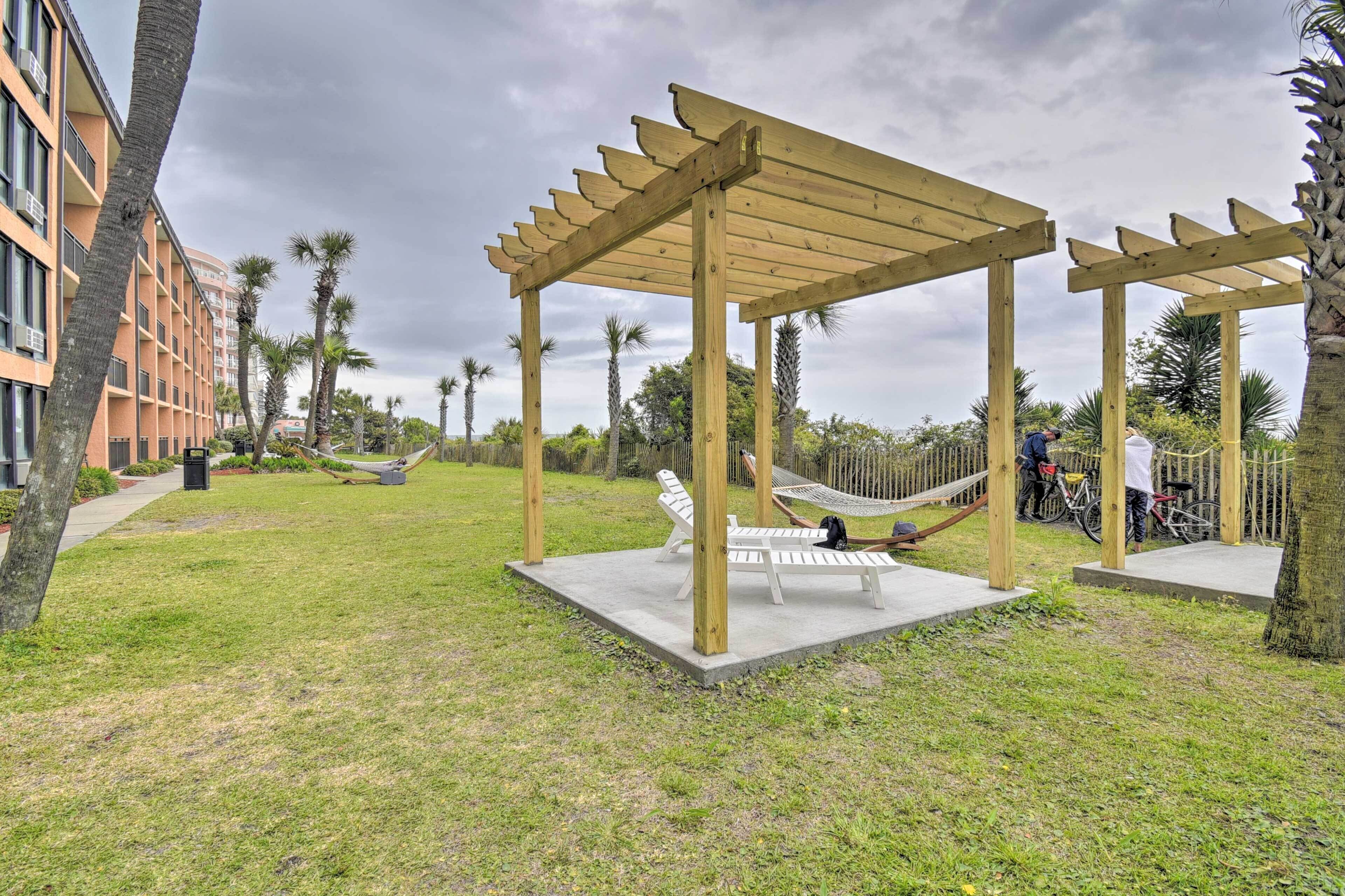 A wooden pergola structure with benches and a hammock in a grassy outdoor area.