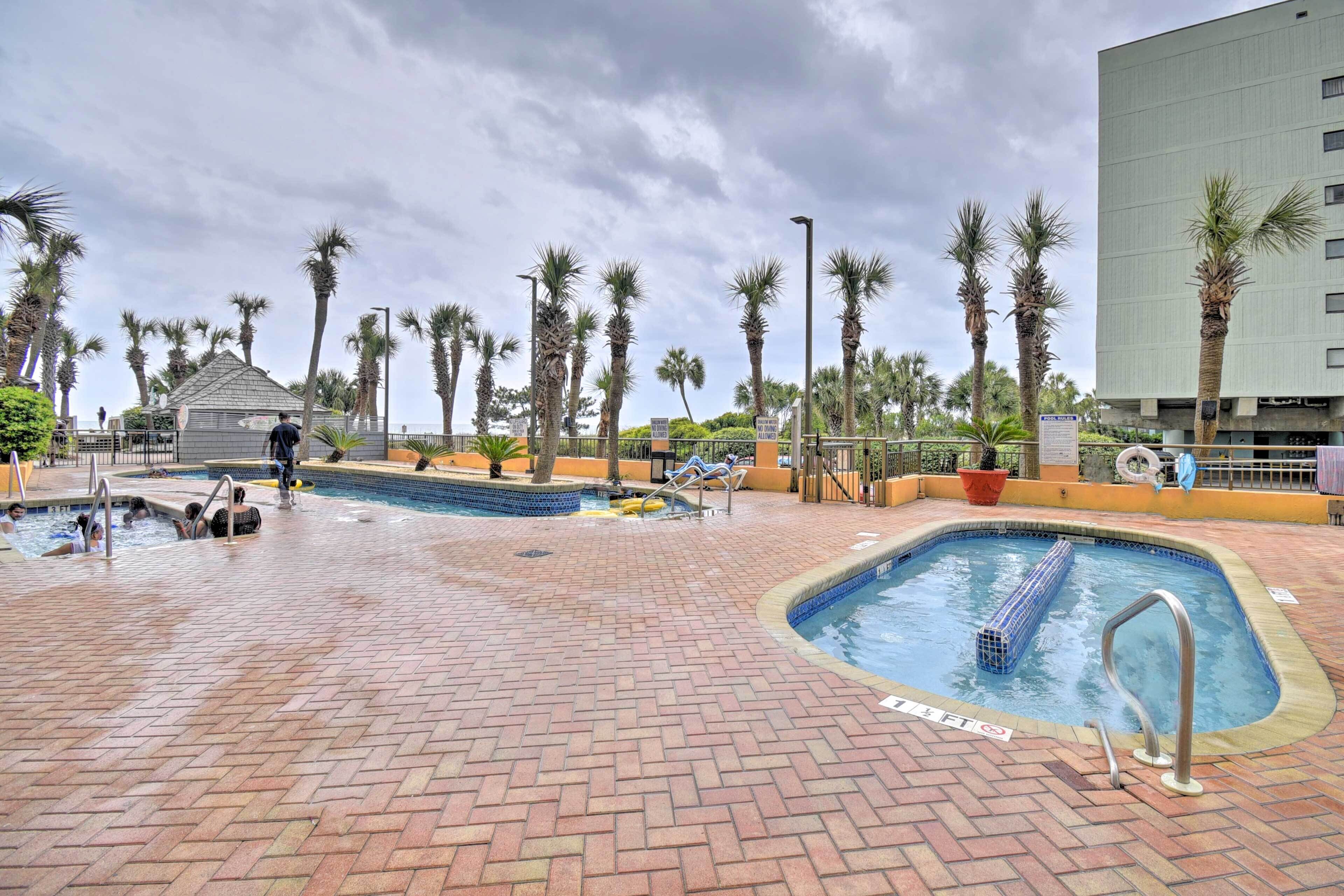 A swimming pool area with multiple pools, palm trees, and a building in the background.