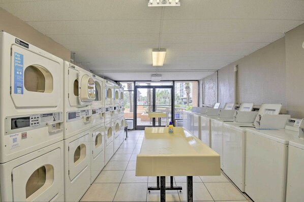 Een wasserette met meerdere wasmachines en drogers, een tafel met een fles wasmiddel en uitzicht op buiten door de glazen deuren.