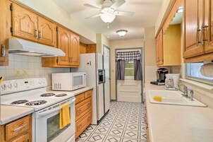 Uma cozinha com armários de madeira, fogão branco, micro-ondas, geladeira, ventilador de teto, piso estampado e janela com cortinas.