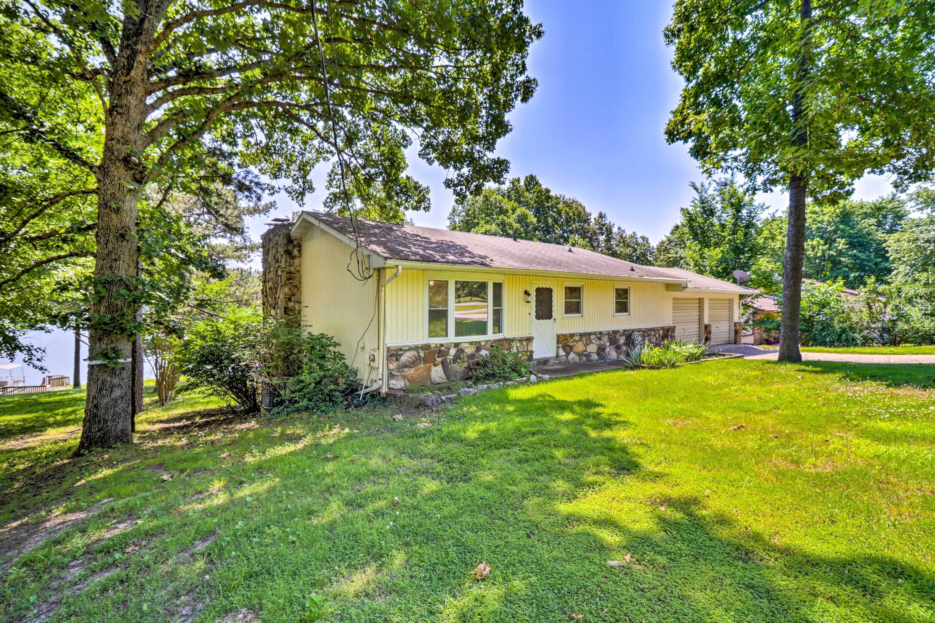 A single-story yellow house with a stone foundation, surrounded by lush green grass and trees.