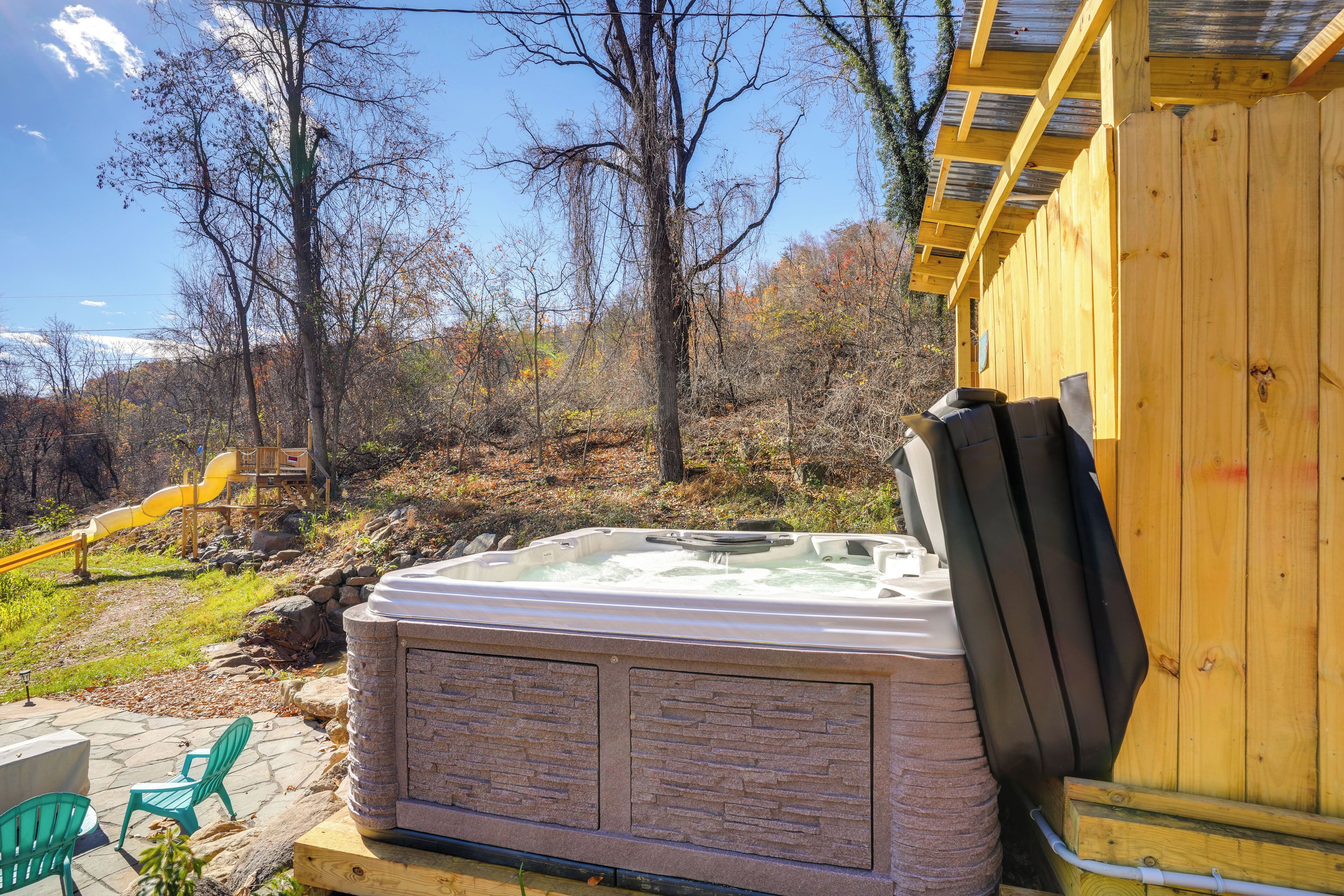 A hot tub with stone panels, a wooden deck, and a slide in a wooded area.