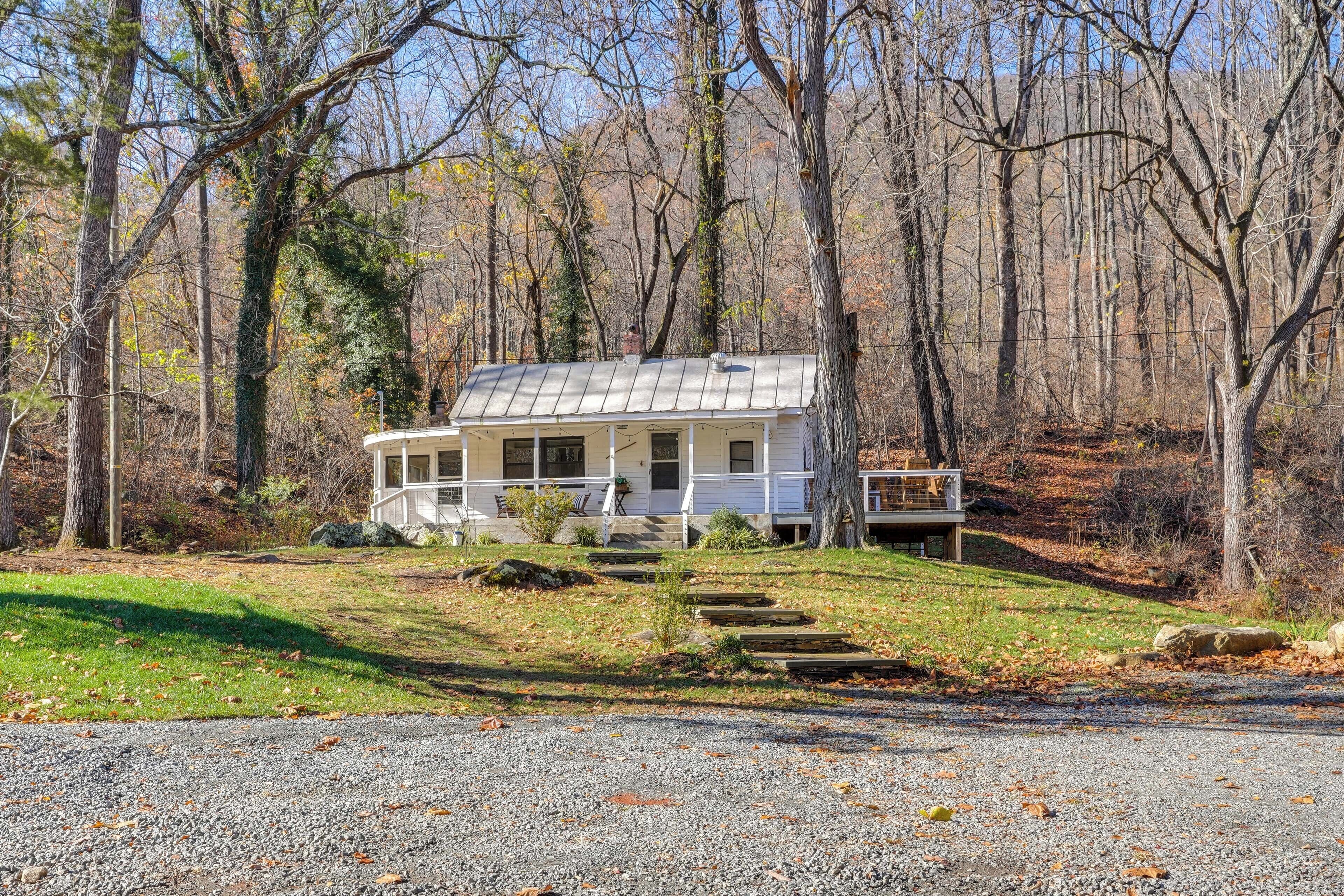 A white cabin with a porch, surrounded by trees and a gravel driveway.
