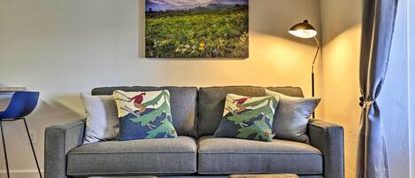 En grå sofa med matchende skammel, et ophængt landskabsmaleri, en gulvlampe og en blå stol.