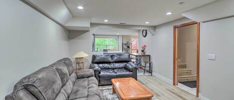 A living room with a sofa, a coffee table, and a rug. There is a kitchen area with a dining table and chairs in the background.