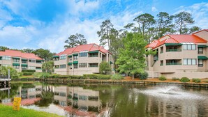 A multi-story building with a red roof, a pond in front, and greenery around.