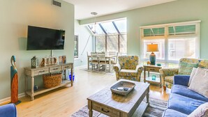 A spacious living room with a flat-screen TV, a wooden coffee table, a blue sofa, and a patterned armchair.