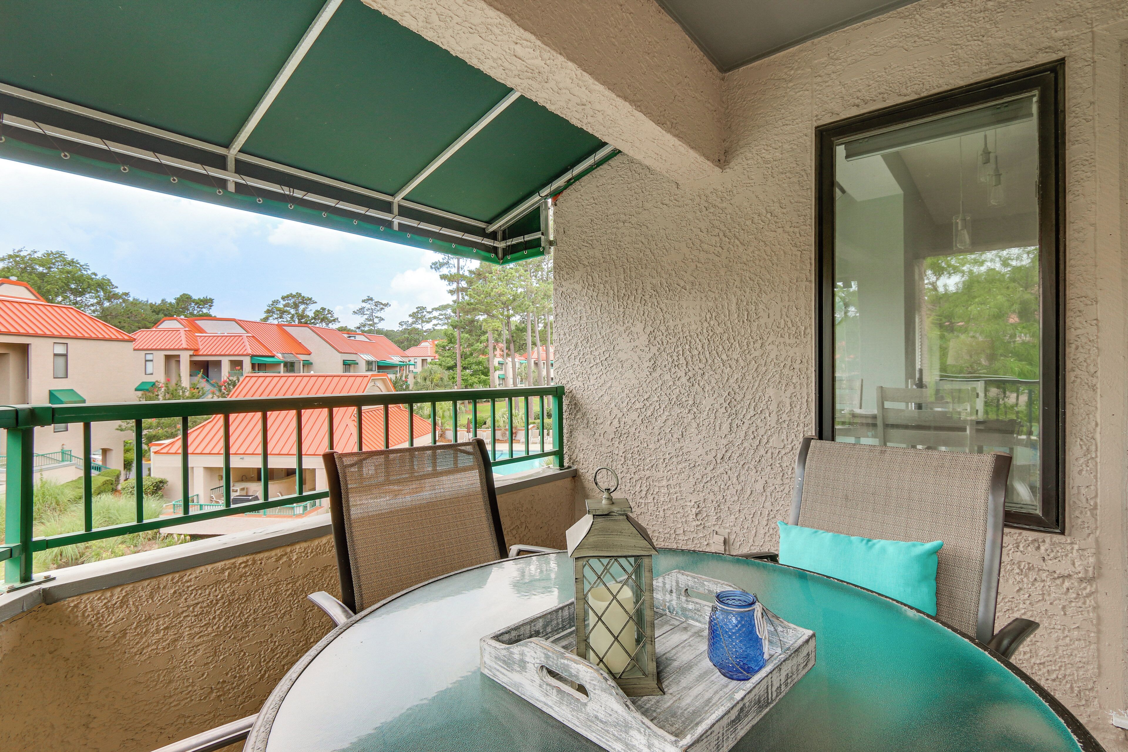 A balcony with a glass table, chairs, a lantern, and a view of buildings.
