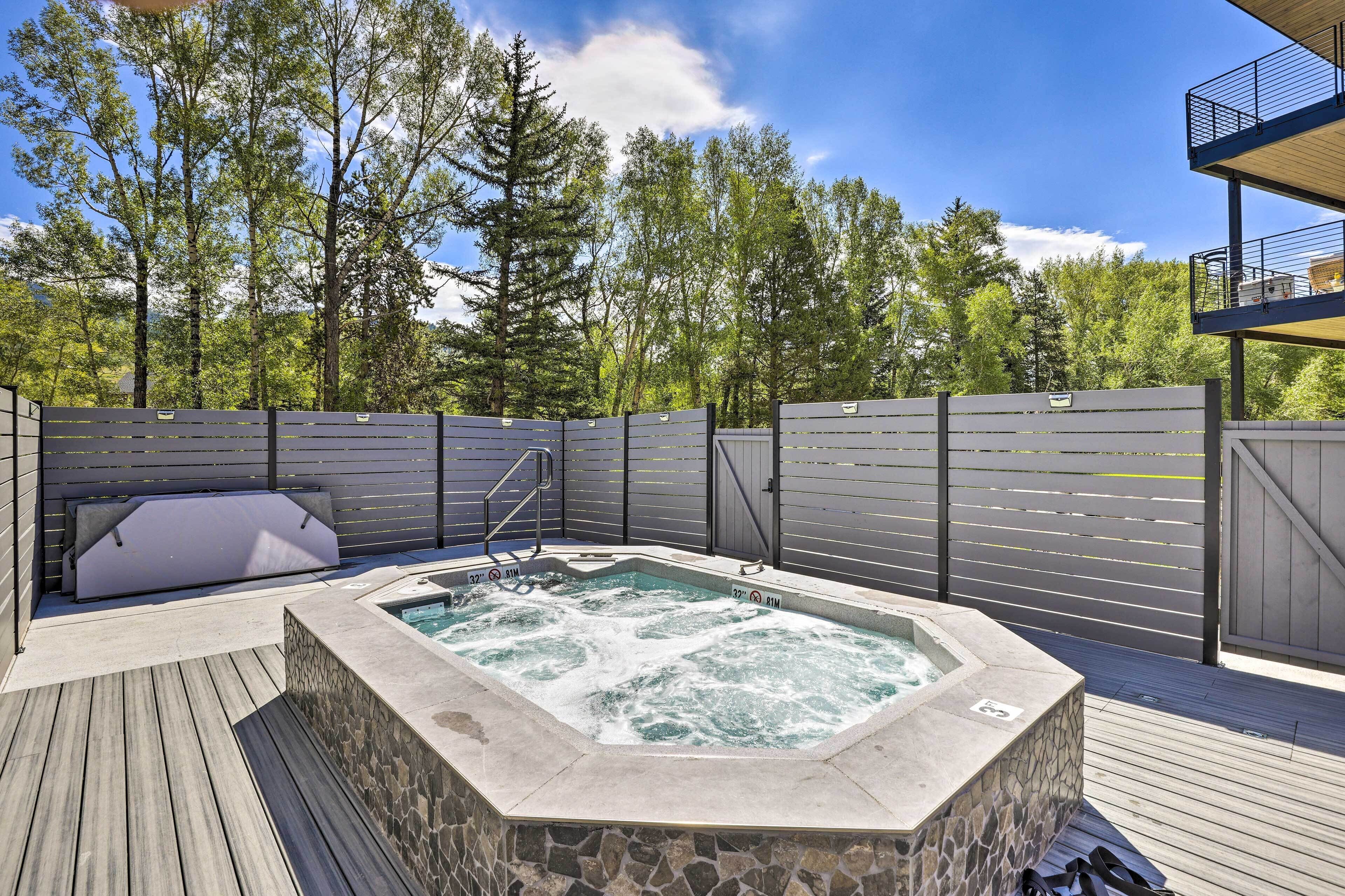 A hot tub on a wooden deck with a stone wall, surrounded by a wooden fence and trees in the background.
