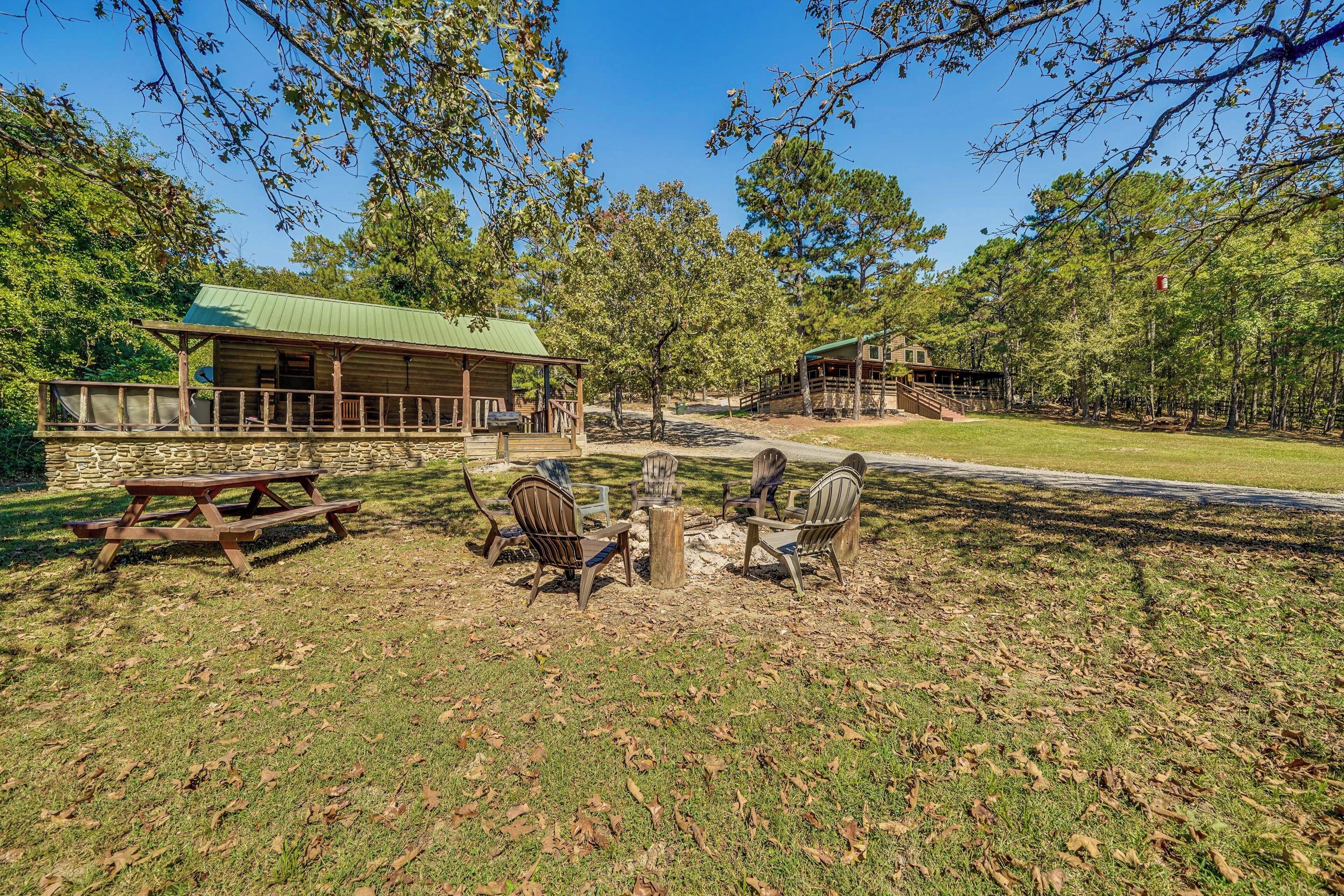 A rustic cabin with a green roof, a picnic table, and three Adirondack chairs in a wooded area.