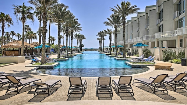 A pool area with lounge chairs and palm trees, surrounded by hotel buildings. - South Padre Island Retreat w/ Beach Access! (South Padre Island)