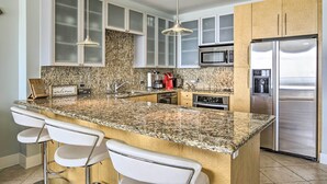 A kitchen with granite countertops, stainless steel appliances, white bar stools, and a granite backsplash. - South Padre Island Retreat w/ Beach Access! (South Padre Island)