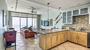 A modern kitchen with a large island, wooden cabinets, and a granite countertop. There is a dining area with a red chair and a sofa. - South Padre Island Retreat w/ Beach Access! (South Padre Island)