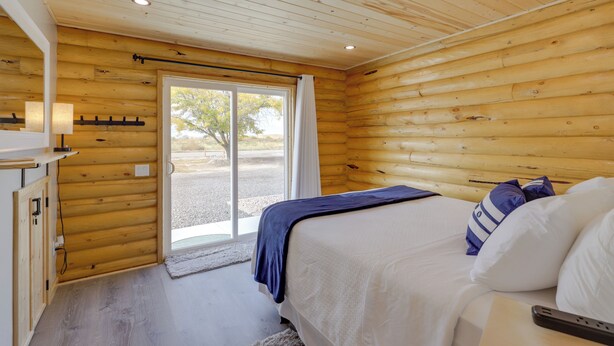 A wooden cabin bedroom with a bed, a nightstand, a lamp, and a window with curtains. - Secluded Grand Junction Getaway w/ Sweeping Views! (Grand Junction)