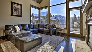 A spacious living room with a large sectional sofa, a fluffy rug, and large windows offering a view of a snowy mountain landscape.