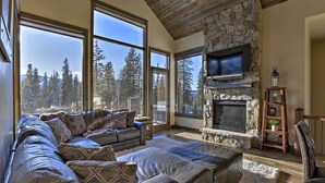 A spacious living room with a stone fireplace, large windows offering a view of trees, and a leather sofa.