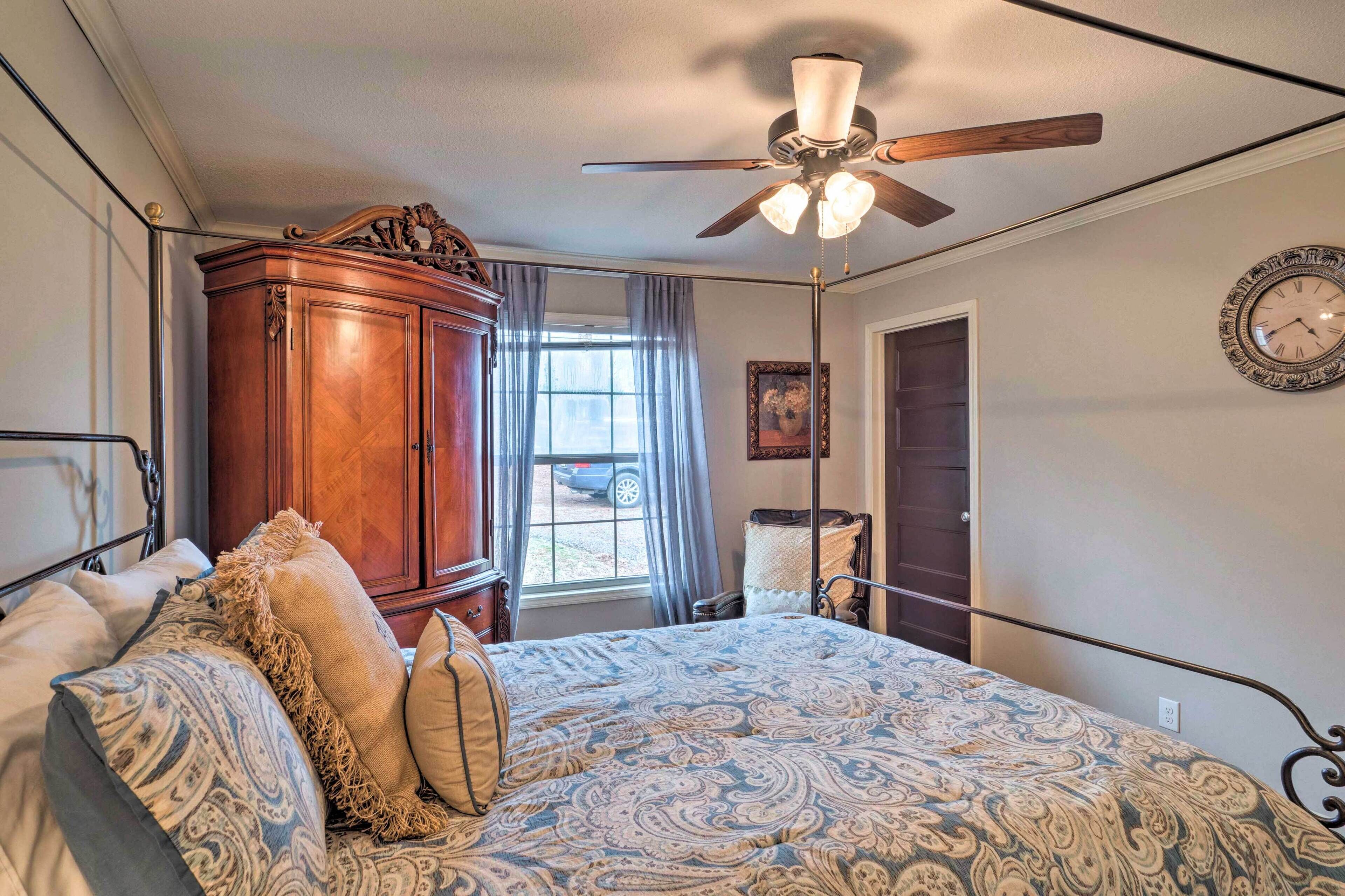 A bedroom with a large bed, a wooden wardrobe, a ceiling fan, a window with curtains, and a wall clock.