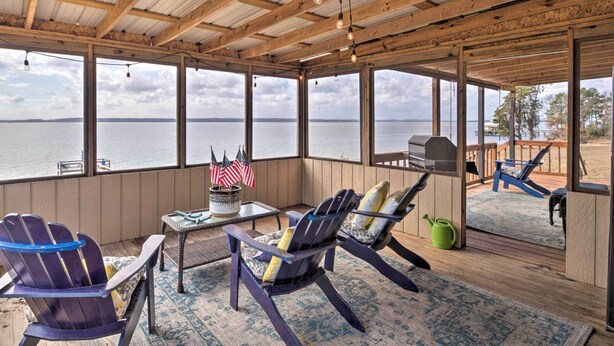 A covered outdoor seating area with Adirondack chairs, a table with an American flag, and a view of the water. - Waterfront Louisiana Vacation Rental w/ Dock! (Many)