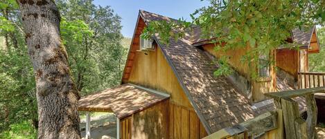A rustic wooden cabin with a sloped roof, surrounded by trees and a wooden deck.