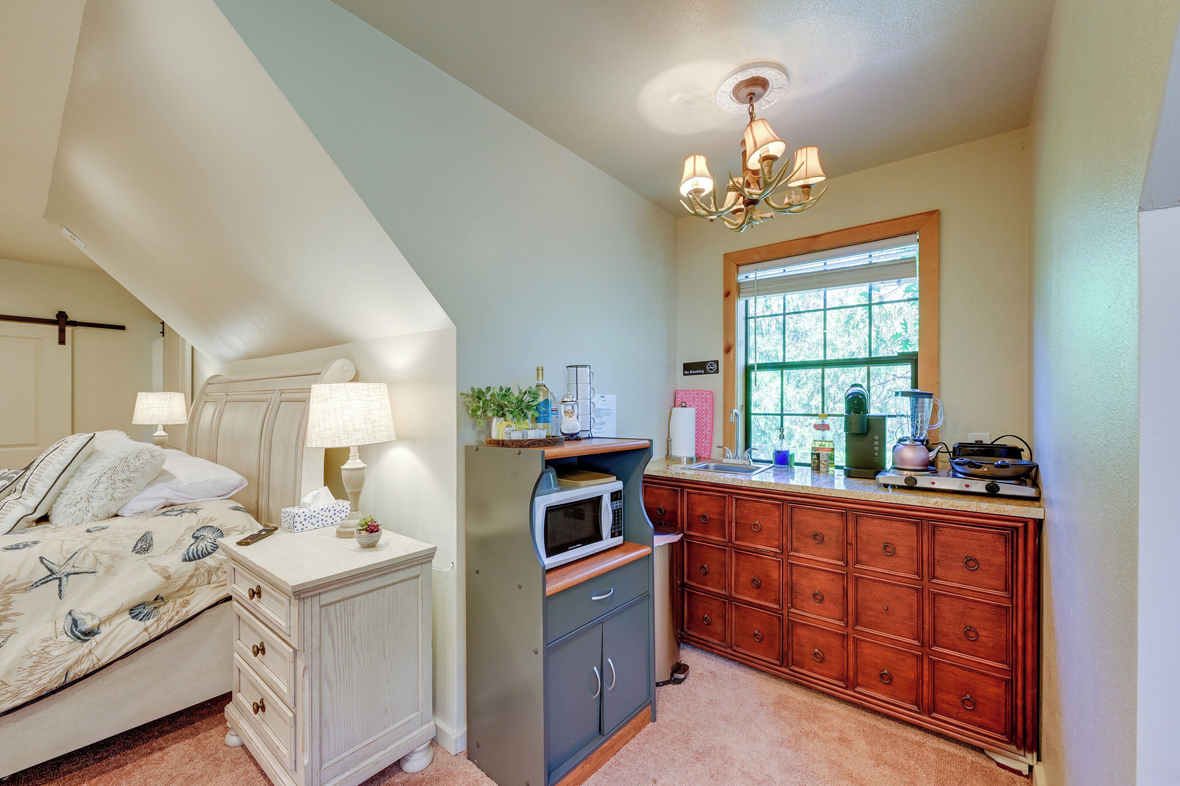 A bedroom with a bed, bedside table, and a nightstand. There is a kitchenette with a microwave, toaster, and coffee maker. A window provides natural light.