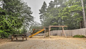 Un parque infantil con tobogán y estructura para escalar, rodeado de árboles y una valla de madera.