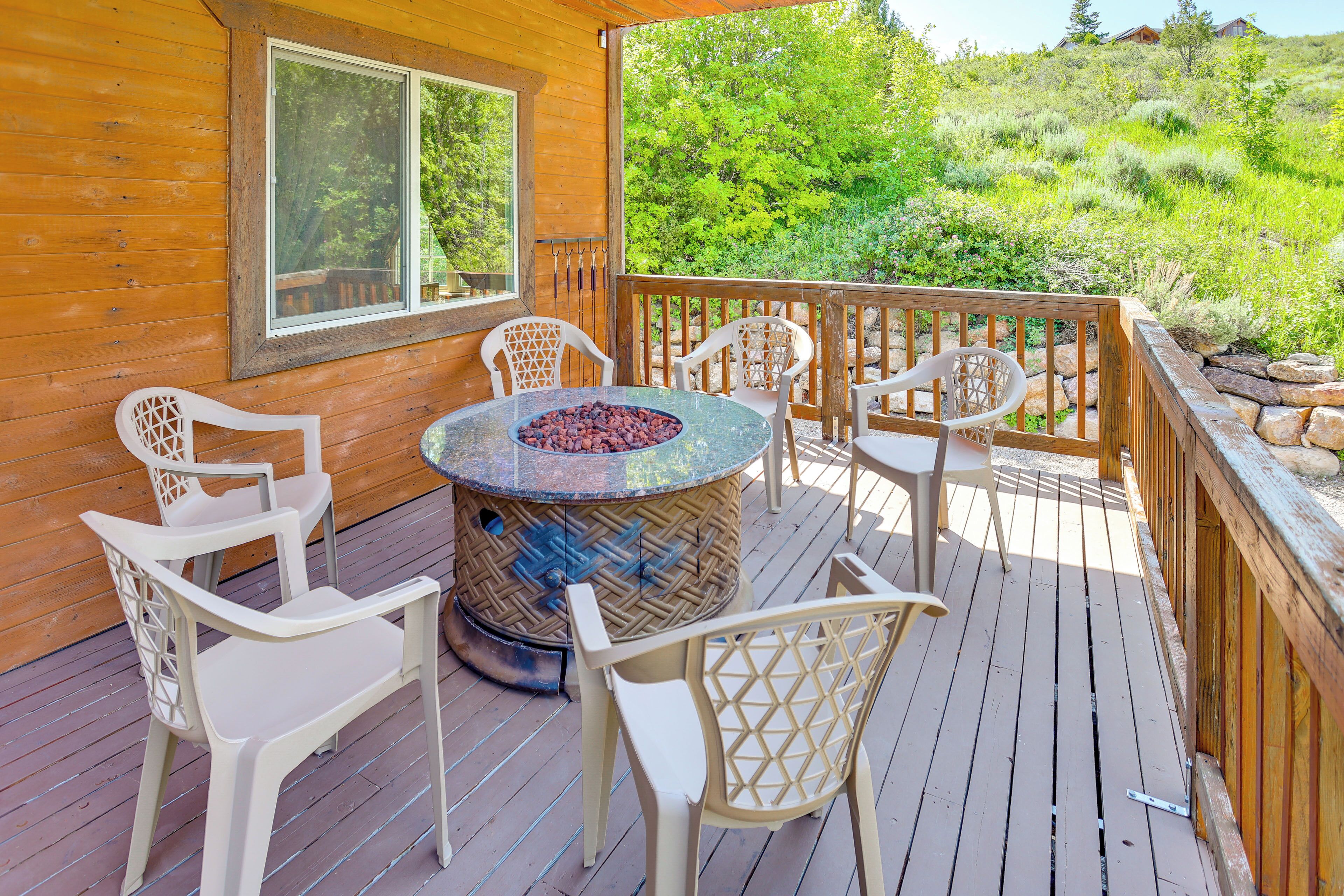 A wooden deck with a round table and chairs, a fire pit, and a view of a green hillside.