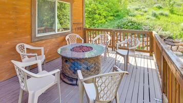 A wooden deck with a round table and chairs, a fire pit, and a view of a green hillside.