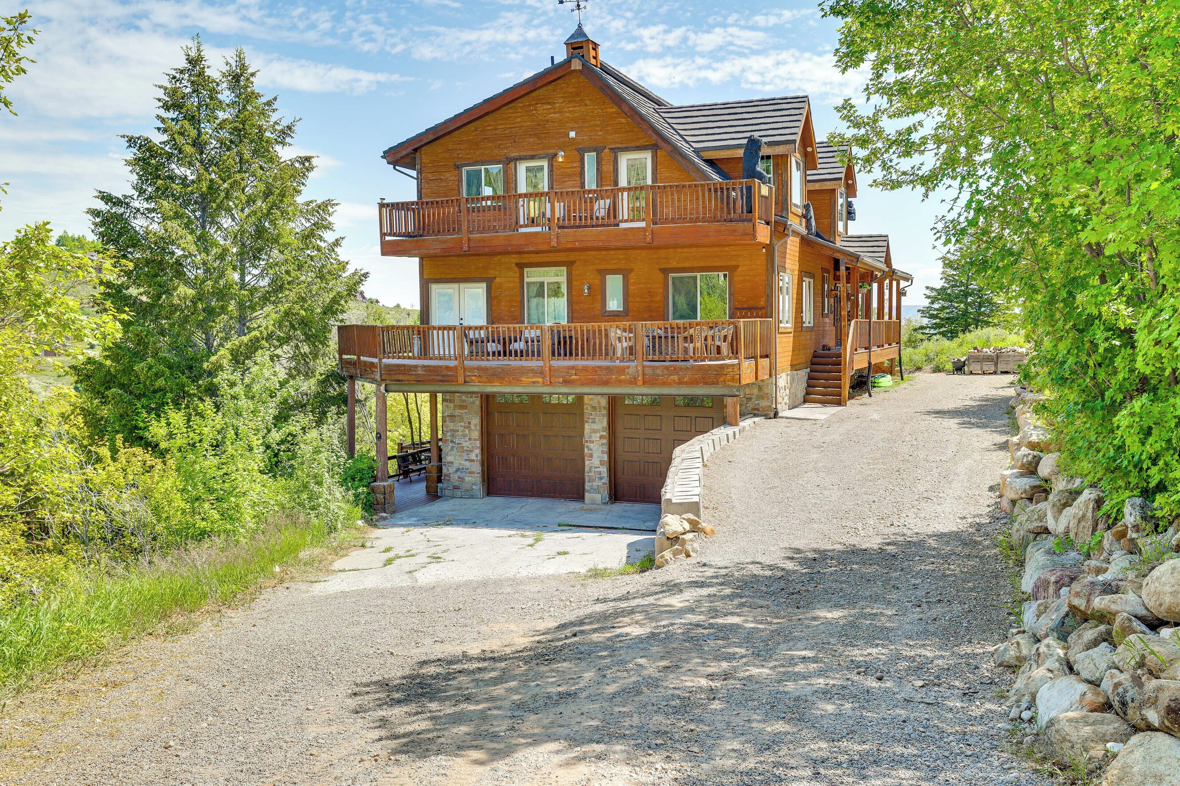 A two-story wooden cabin with a balcony, a gravel driveway, and a stone wall on one side.