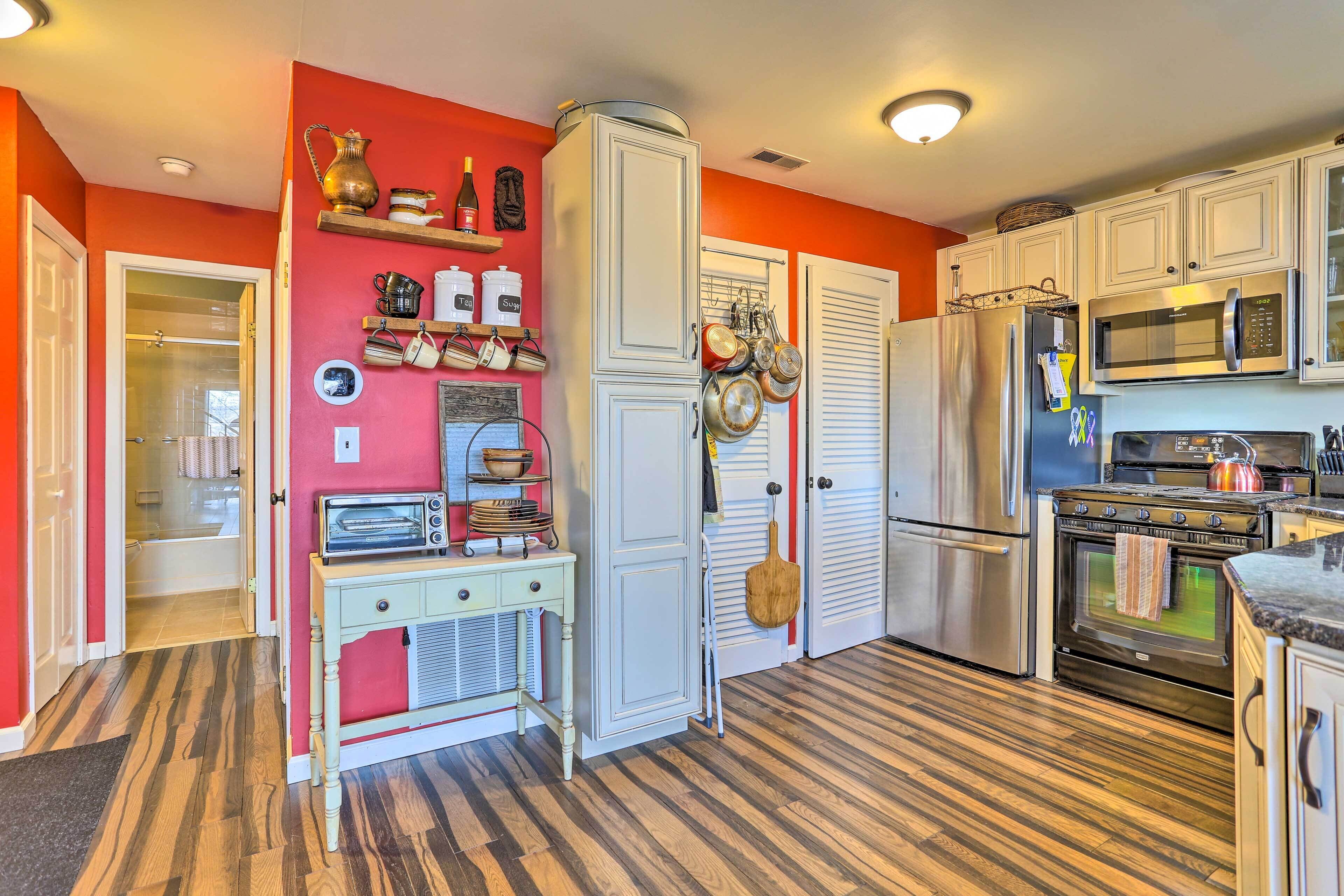Una cocina con armarios blancos, microondas, estufa y refrigerador. Hay una mesa de madera con una radio y una repisa con utensilios de cocina.
