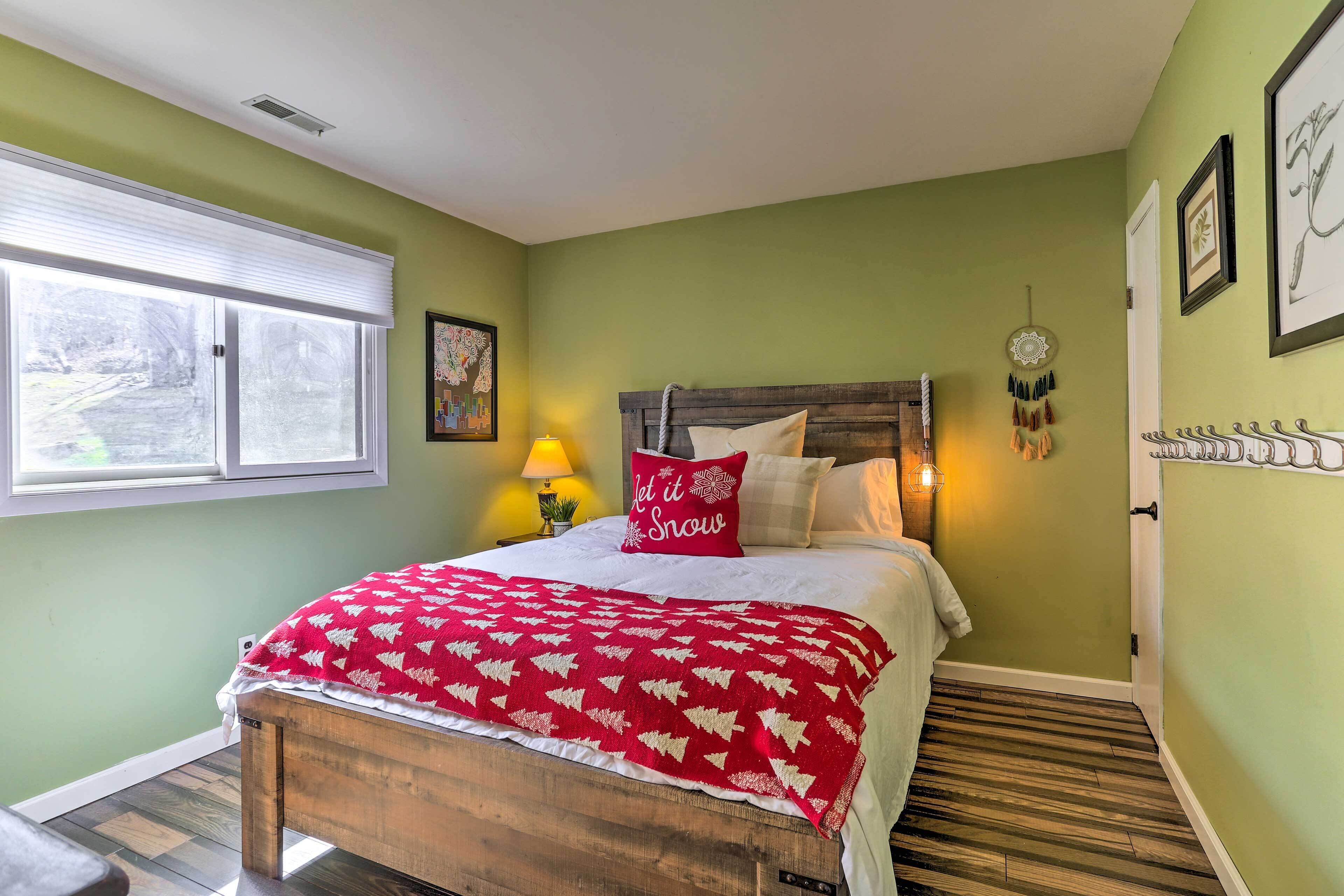 A bedroom with a wooden bed, a red and white patterned blanket, a pillow with "Let it Snow" text, a wall-mounted dreamcatcher, and a window with blinds.
