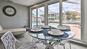Une table en verre, quatre assiettes et bols blancs, une chaise blanche, une horloge au mur et une vue sur une piscine et des bâtiments à l’extérieur.