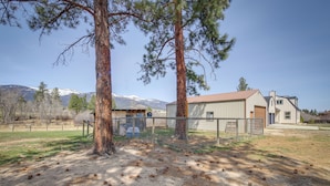 A fenced outdoor area with pine trees, a building with a red roof, and mountains in the background. - Pet-friendly Victor Home w/ Fire Pit & Large Yard! (Victor)