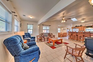 A spacious living area with a blue armchair, a wooden rocking chair, a coffee table, and a sofa. There are multiple windows providing natural light, and a kitchen area with wooden cabinets and a refrigerator is visible in the background.