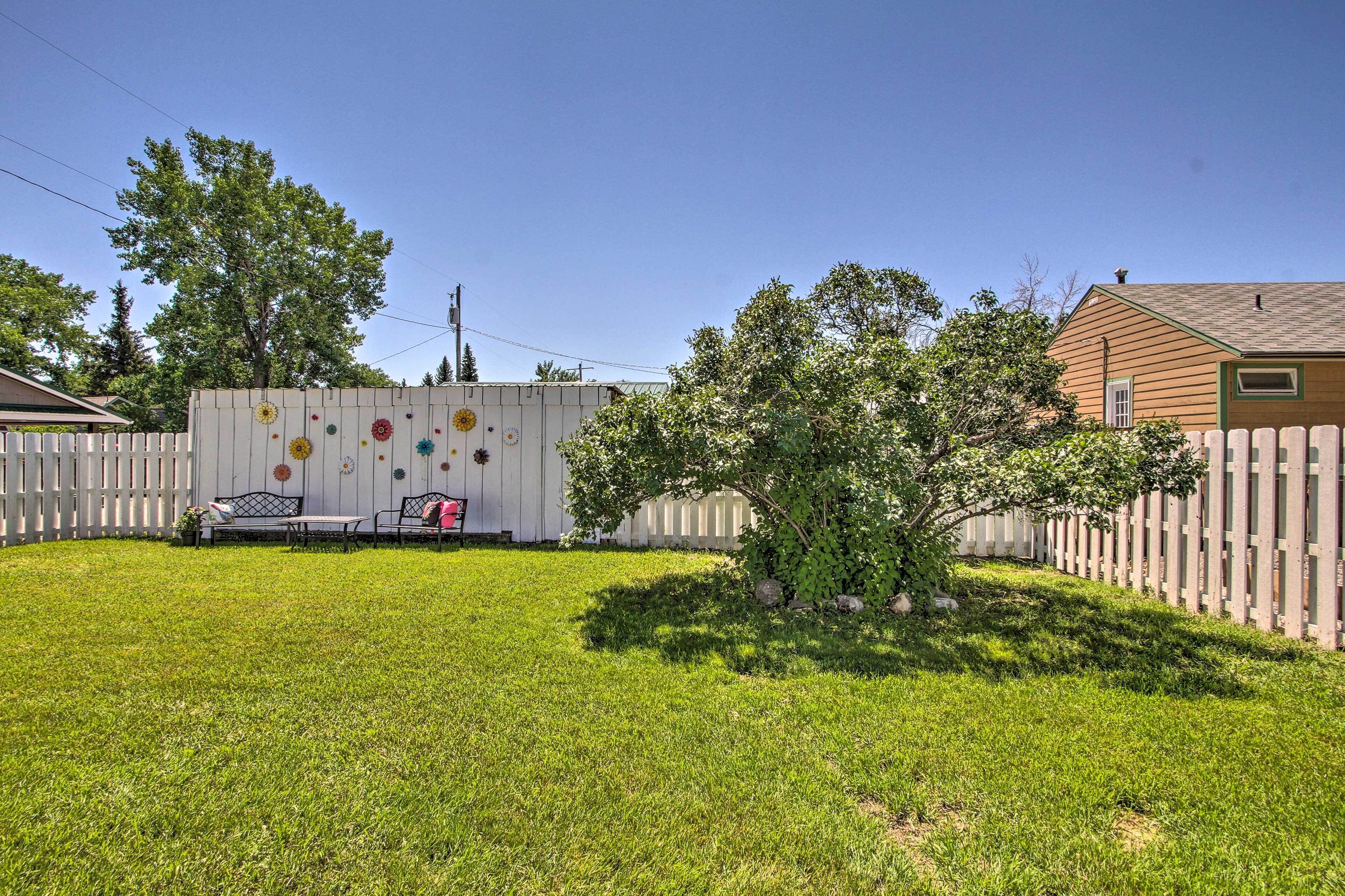 A backyard with a white fence, a small tree, and a patio area with a table and chairs.
