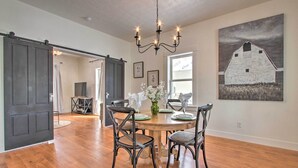 A dining area with a wooden table, four chairs, a chandelier, and a painting of a barn on the wall. - Charming Choteau Cottage: Ski & Fish Nearby! (Choteau)