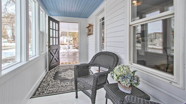 A covered porch with a wicker chair and a table, a door leading outside, and a window with a view of the outside. - Charming Choteau Cottage: Ski & Fish Nearby! (Choteau)