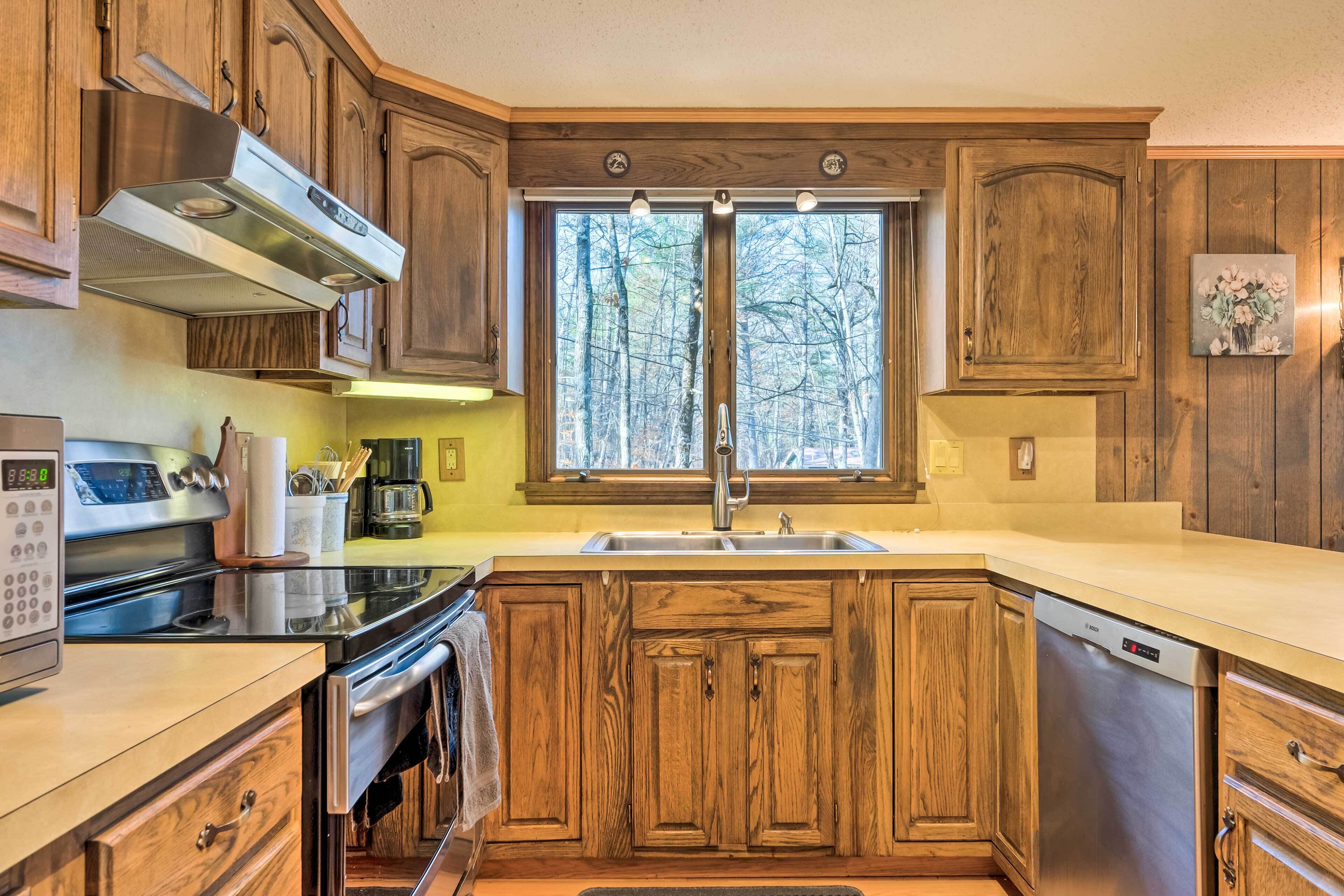 A kitchen with wooden cabinets, a stainless steel range hood, a double sink, and a window with a view of trees.