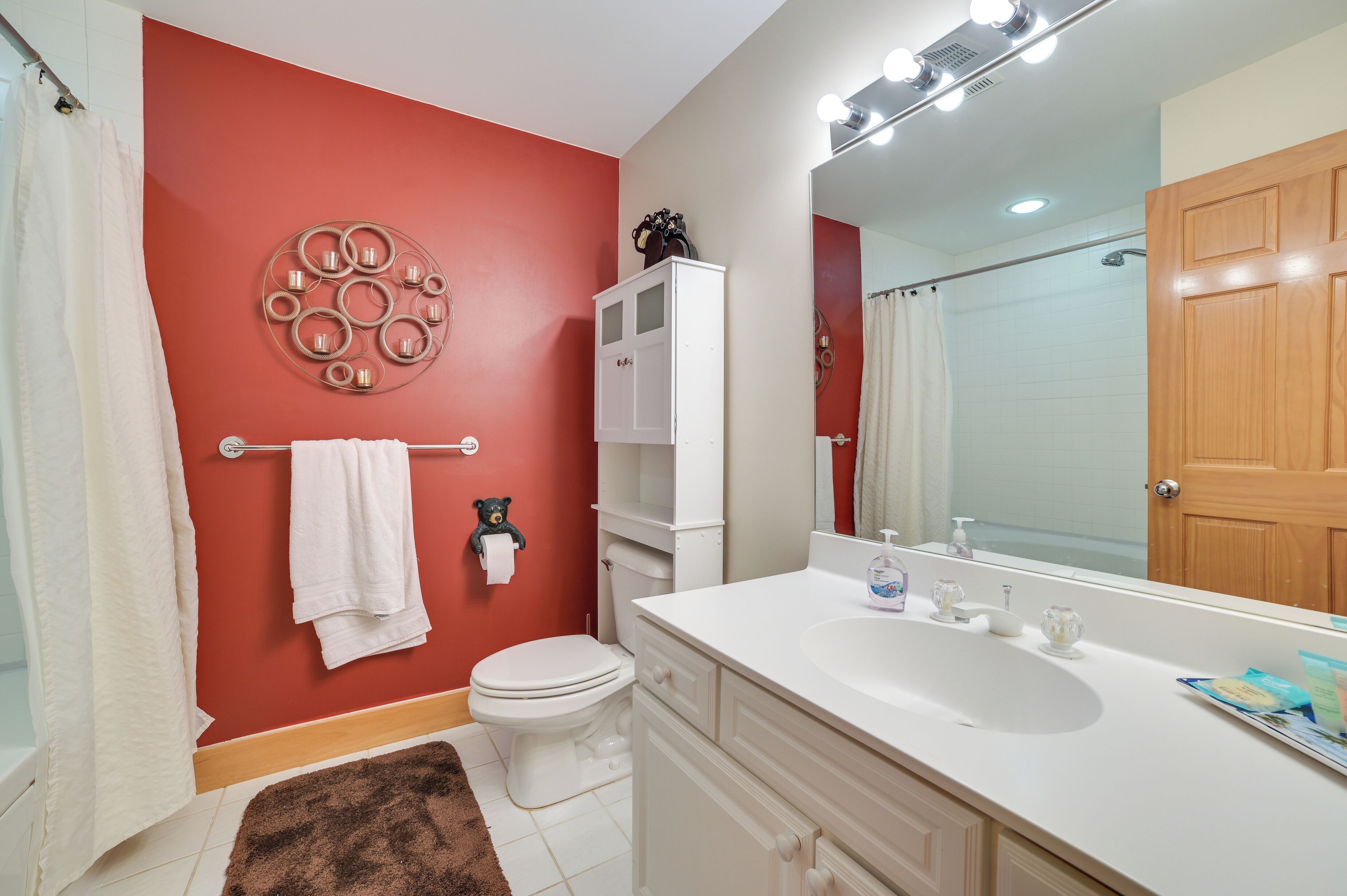 A bathroom with a red wall, white fixtures, a large mirror, and a wooden door.