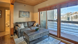 A living room with a wooden ceiling, a gray sofa with cushions, a coffee table, and a large sliding glass door leading to a balcony with a view of buildings. - Cozy Crested Butte Condo - Walk to Ski Lift! (Crested Butte)