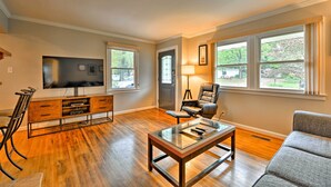 A spacious living room with hardwood flooring, a glass coffee table, a leather armchair, a sofa, a TV mounted on the wall, and a wooden entertainment unit.