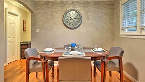 A dining room with a round table set for four, a wall clock, and a window with blinds.