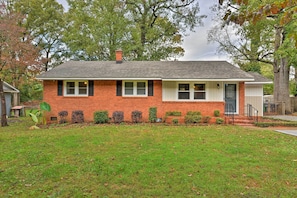 A single-story brick house with a front porch, a small front yard, and a tree in the background.