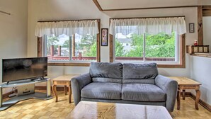 A living room with a grey sofa, a TV on a stand, two windows with curtains, and a wooden floor. - Charming Laconia Home w/ Deck: Walk to Lake! (Laconia)