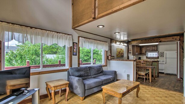 A living room with a gray sofa, wooden coffee table, and a TV. There are windows with white curtains, a kitchen area with a refrigerator, stove, and microwave. - Charming Laconia Home w/ Deck: Walk to Lake! (Laconia)