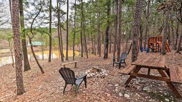A picnic area with a wooden table and chairs, surrounded by trees and a small fire pit.